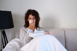 Osoba siedząca na sofie pod białą kołdrą, trzymająca pudełko chusteczek