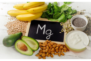 Produkty spożywcze bogate w magnez ułożone wokół tabliczki z napisem „Mg”, w tym banany, awokado, szpinak, migdały, nasiona i jogurt, przedstawiające dietę wspierającą uzupełnienie niedoboru magnezu.