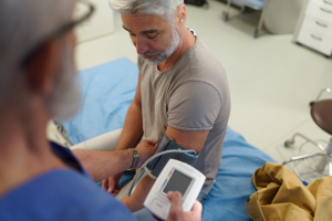 Senior siedzący na łóżku w gabinecie medycznym, podczas pomiaru ciśnienia krwi mankietem założonym na ramię przez lekarza