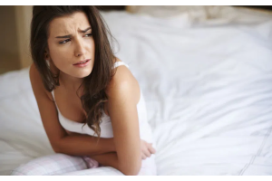 Osoba siedząca na łóżku w jasnym pomieszczeniu, ubrana w białą koszulkę, trzymająca ręce na brzuchu