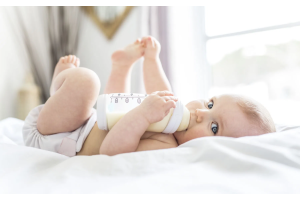 Niemowlę leżące na jasnym łóżku, trzymające w dłoniach butelkę z mlekiem i pijące z niej, ubrane w jasną pieluszkę, w tle widoczne wnętrze pokoju z oknem.