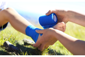 Zbliżenie na stopę ułożoną na trawie, druga osoba owija staw skokowy niebieskim bandażem elastycznym, dłonie trzymają rolkę opaski