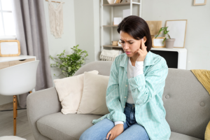 Osoba siedząca na sofie w domowym wnętrzu, ubrana w zieloną koszulę i jeansy, trzymająca rękę przy uch