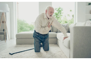 Osoba klęcząca na podłodze w jasnym salonie, jedną dłonią opierająca się o sofę, drugą trzymająca się za klatkę piersiową