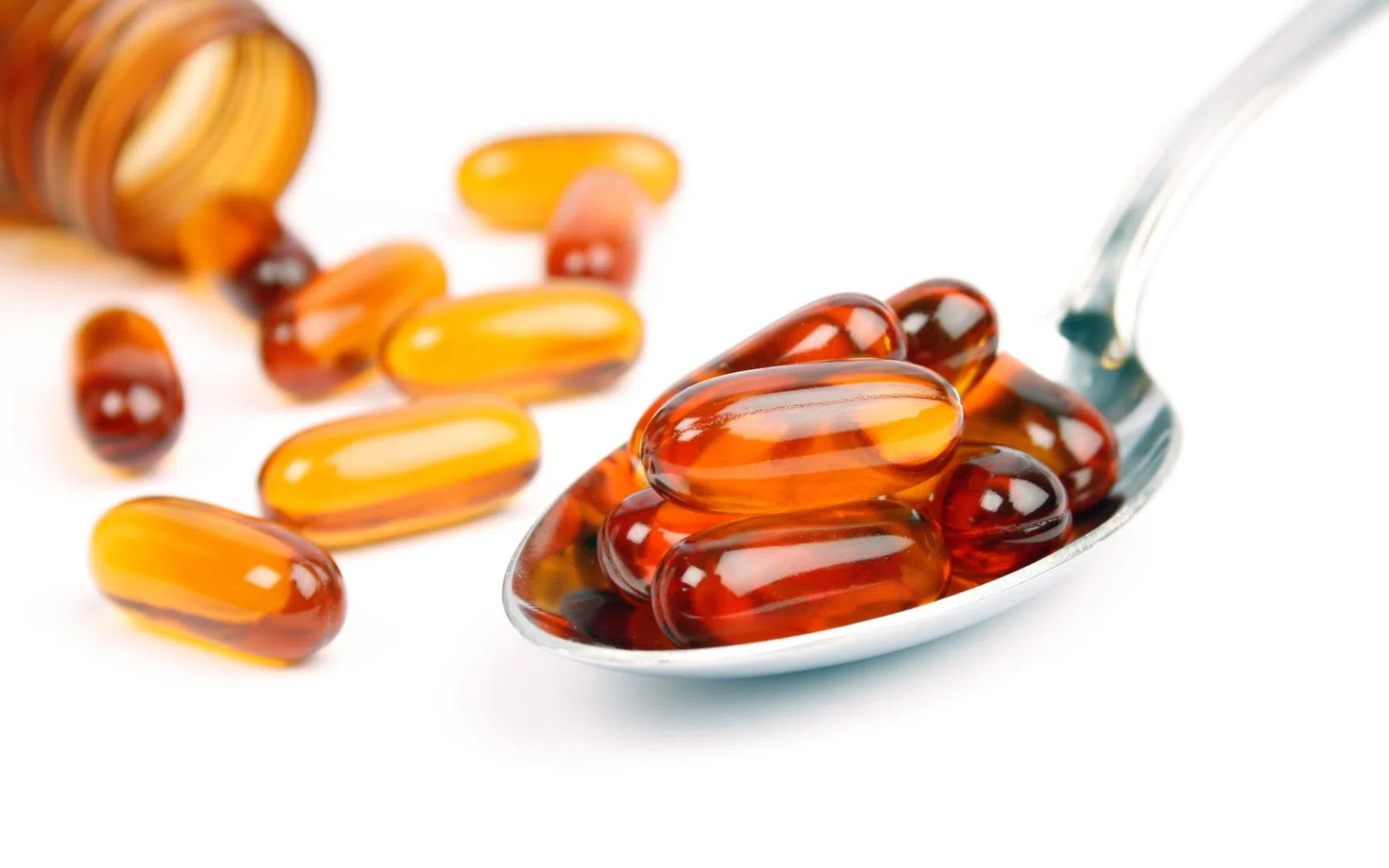 Przezroczyste kapsułki z bursztynową zawartością leżące na łyżce i wysypane z brązowej butelki na białym tle