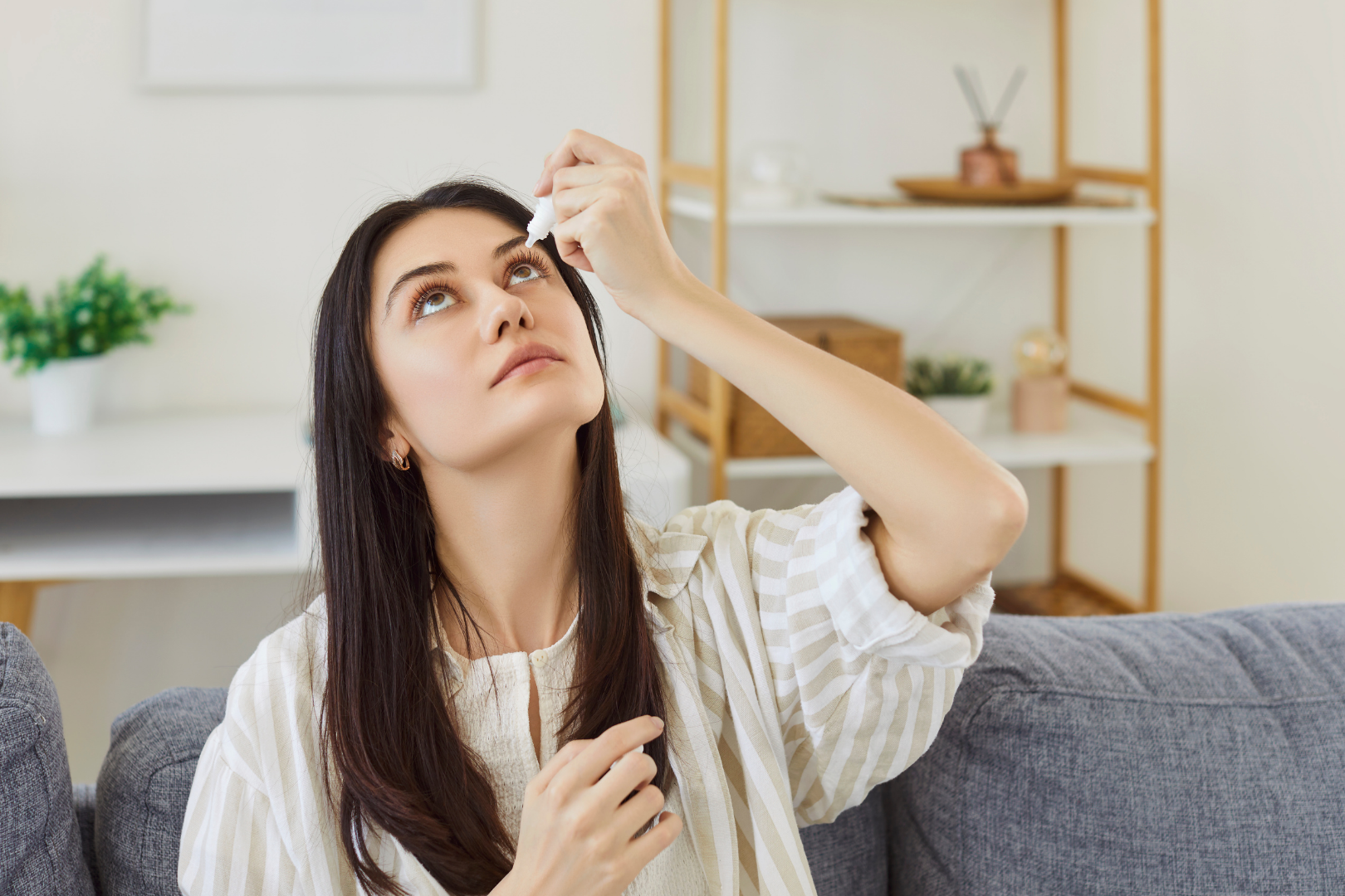 Osoba siedząca na kanapie w jasnym wnętrzu aplikuje krople do oka z małej butelki, głowa lekko uniesiona, dłoń trzyma zakraplacz
