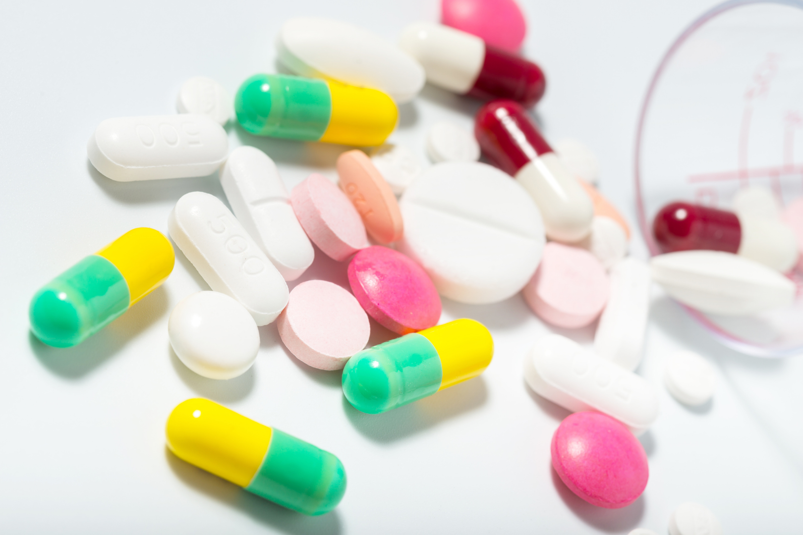 Różnokolorowe tabletki i kapsułki rozsypane na białym tle, w tym żółto‑zielone kapsułki, białe tabletki i różowe drażetki – różne formy leków i suplementów.
