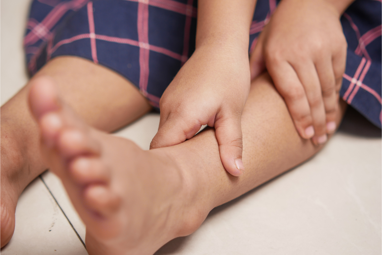Dziecko siedzące na podłodze, trzymające dłonią okolice podudzia i kostki, ujęcie z bliska na nogi i ręce