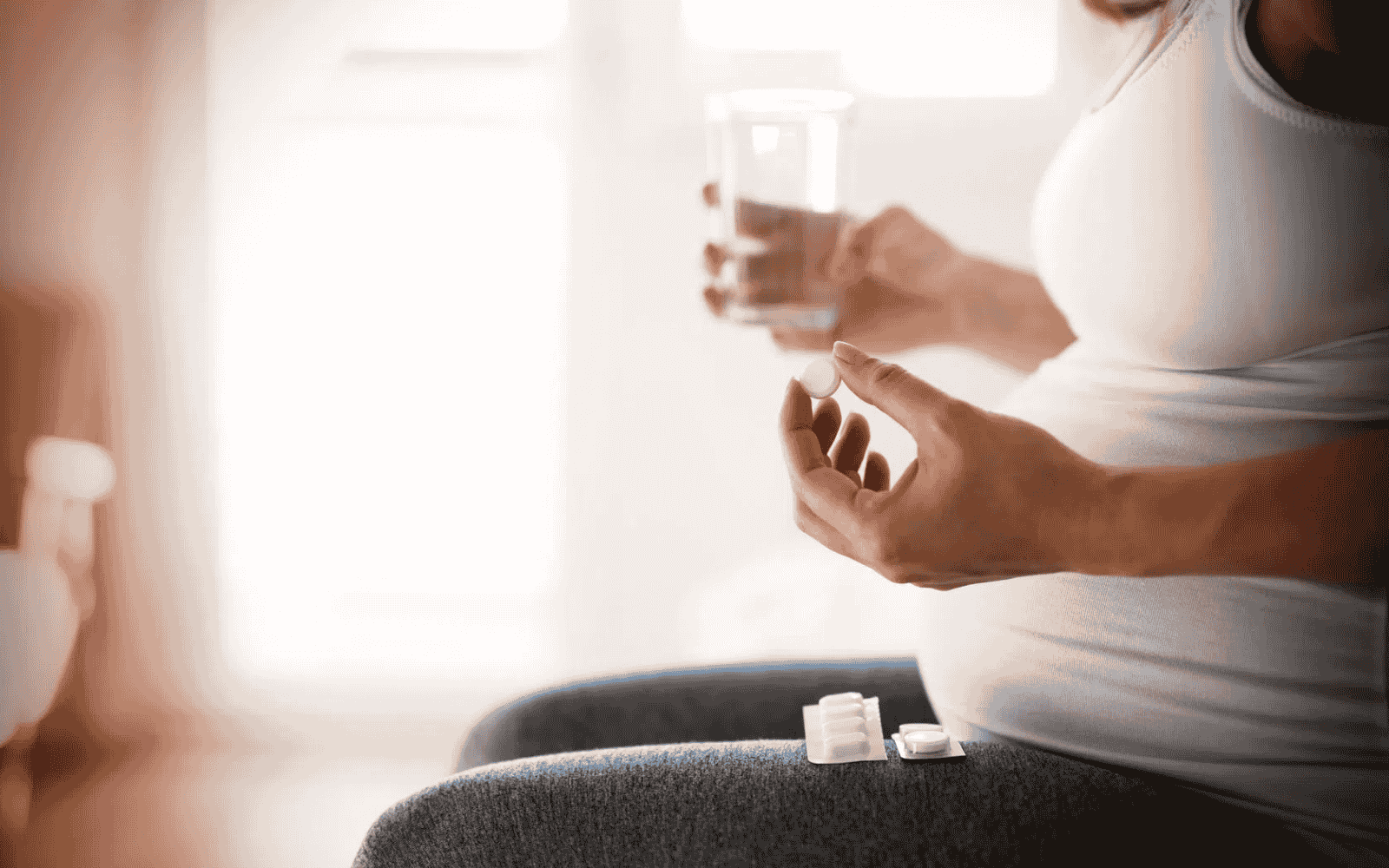 Osoba w ciąży siedząca na kanapie, ubrana w jasny top, trzymająca w jednej dłoni szklankę wody, a w drugiej tabletkę, na kolanach opakowanie leków