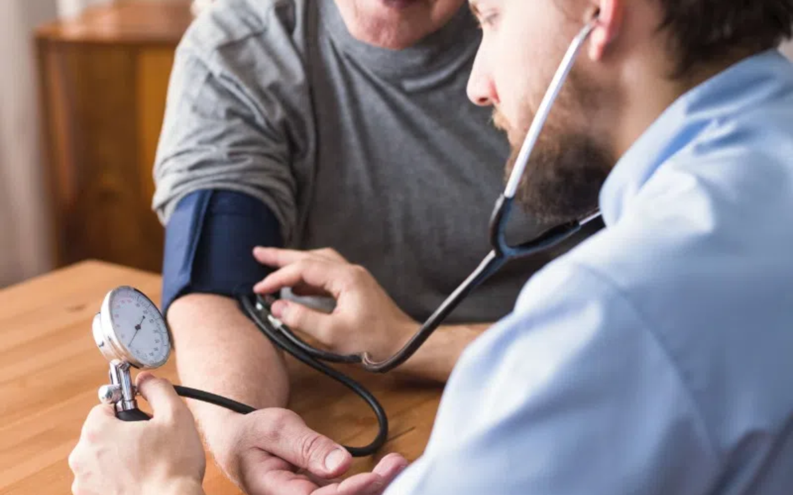 Pomiar ciśnienia krwi wykonywany w domu lub gabinecie – osoba siedząca przy stole z mankietem ciśnieniomierza na ramieniu, druga osoba kontroluje wynik za pomocą aparatu i stetoskopu.