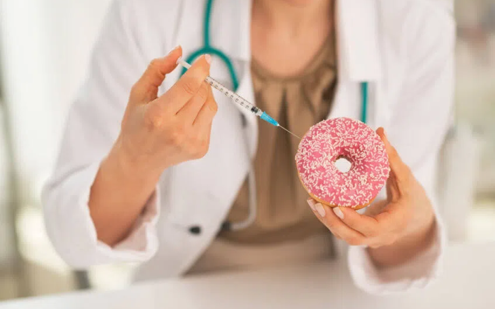 Dłonie osoby w białym fartuchu trzymające różowego pączka posypanego cukrem oraz strzykawkę skierowaną w jego stronę
