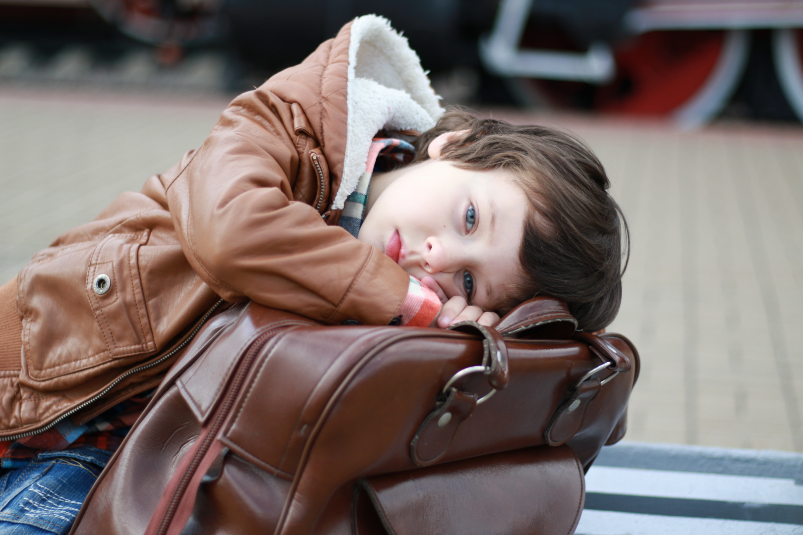 Dziecko leżące na torbie podróżnej w przestrzeni publicznej, ubrane w kurtkę, z głową opartą na bagażu