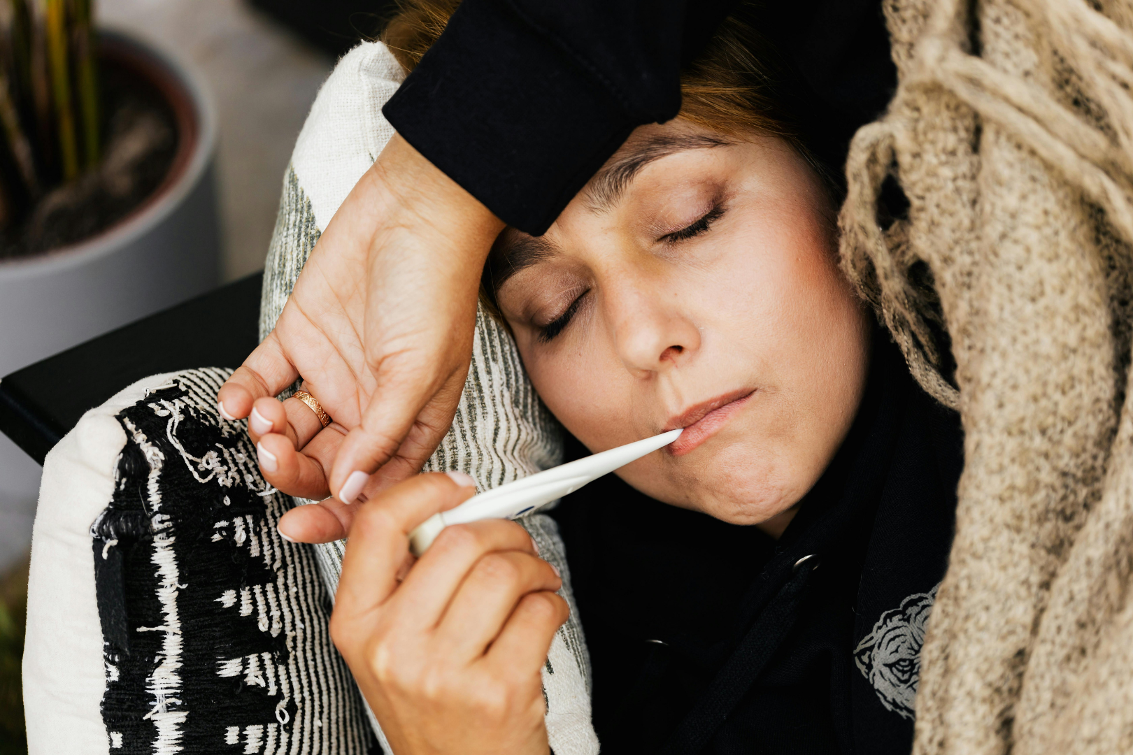 Osoba leżąca pod kocem mierzy temperaturę termometrem i przykłada dłoń do czoła, obraz przedstawiający objawy ospy wietrznej u dorosłych, takie jak gorączka, osłabienie i złe samopoczucie.