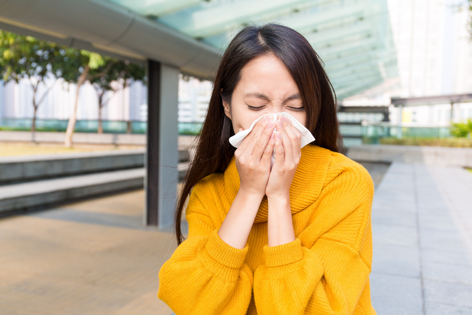 Osoba stojąca na zewnątrz pod zadaszeniem, ubrana w żółty sweter, trzymająca dłonie przy nosie i ustach