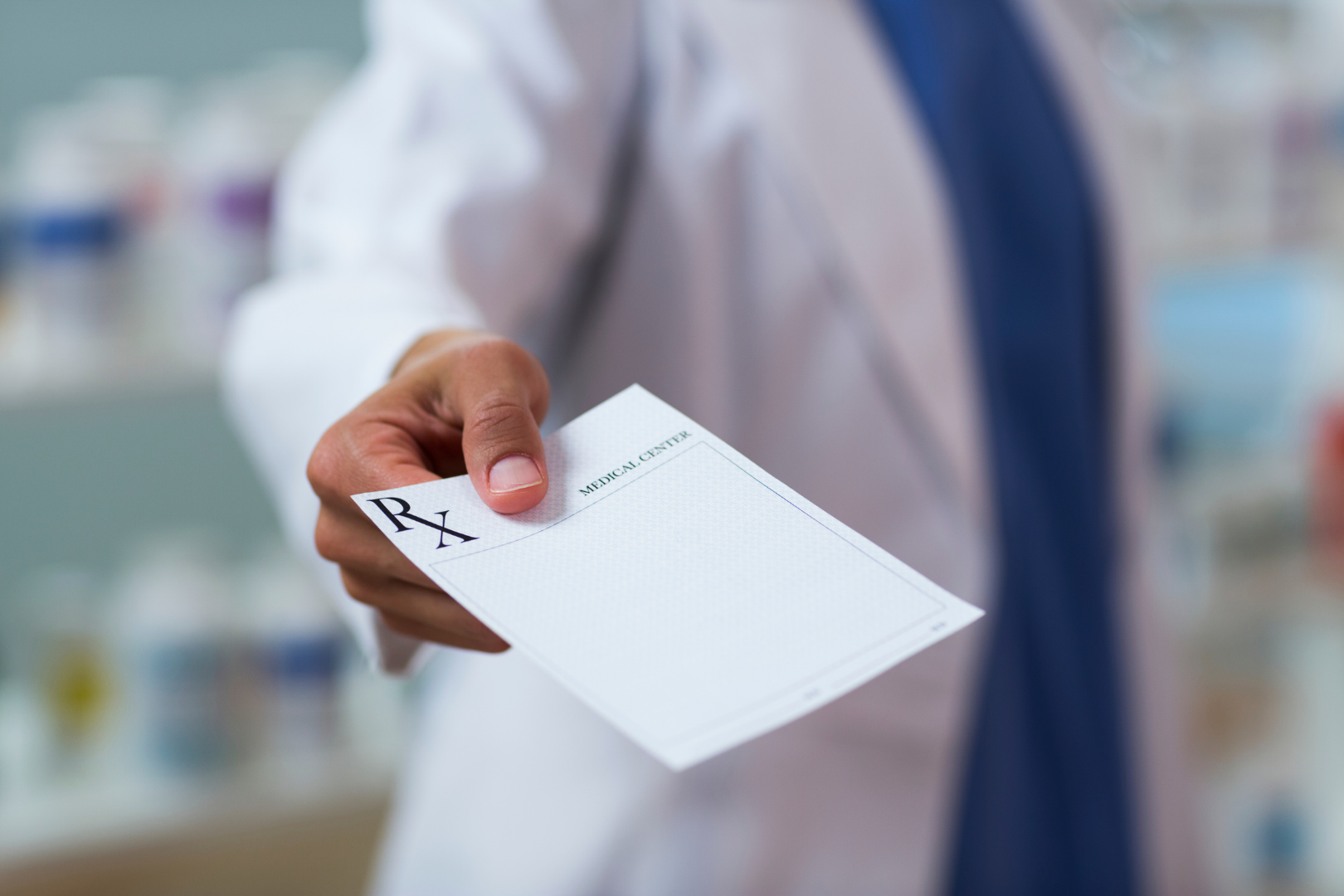 Dłoń wyciągnięta w stronę obiektywu trzymająca druk recepty z oznaczeniem Rx, w tle wnętrze apteki