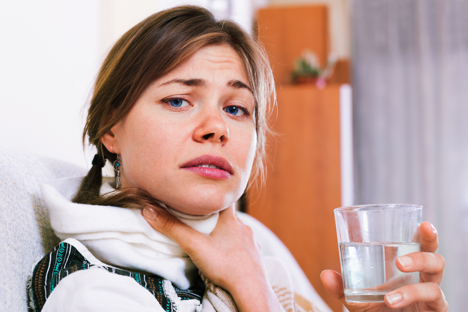 Osoba siedząca w domowym wnętrzu, owinięta białym szalem, trzymająca szklankę wody w jednej dłoni i drugą rękę na szyi 